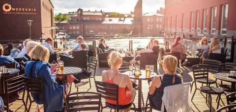 Tamperelaisia Tammerkosken rannalla olevalla terassilla kesäiltana. Etualalla on naisia selin katsojaan. Aurinko paistaa.