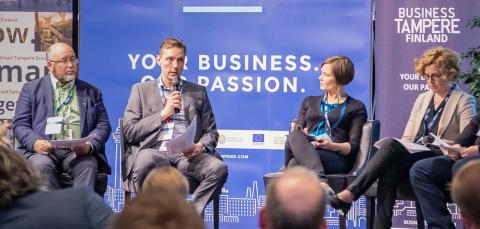 Seitsemän henkilöä paneelikeskustelussa lavalla, yksi puhuu mikrofoniin