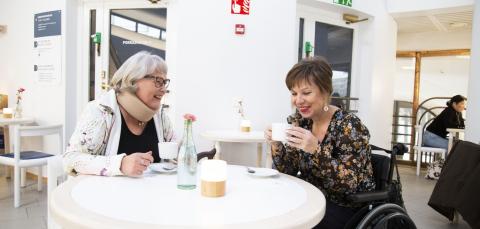 Naiset istuvat ja juovat kahvia pöydän ääressä. Toinen naisista istuu pyörätuolissa.