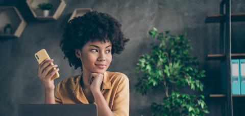 Nuori kiharahiuksinen nainen istuu tummassa toimistossa. Hän pitelee kännykkää kädessään ja katsoo mietteliäänä sivulle. Hänen edessään on kannettava tietokone.