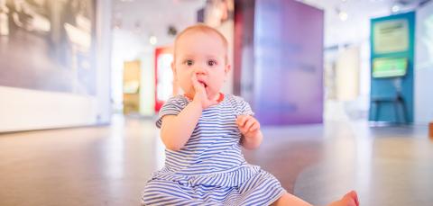 Vauva istuu museonäyttelyn lattialla raidallisessa mekossa.