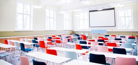 The bright lecture hall has rows of white tables and colourful chairs with velvet cushions. The back wall of the room is covered with a screen.