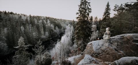 Tupsulakkinen ihminen istuu korkealla kalliolla ja katselee alkutalven lumista metsää.