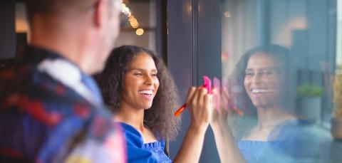 A smiling woman writes on a pink sticky note attached to a glass wall, while a person next to her observes.