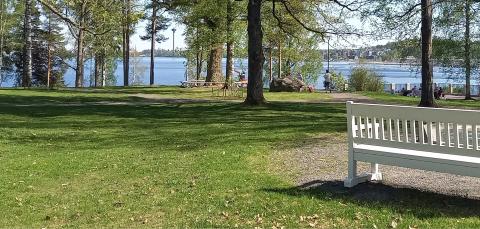 Hiedanrannan kartanon piha kesäisenä päivänä. Etualalla puistonpenkki suihkulähteen vierellä. Taustalla ihmisiä viettämässä aikaa. Pihassa kastaa puita ja Näsijärvi on näkyy taustalla.