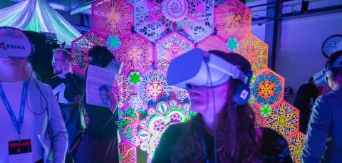 Kaksi henkilöä VR-lasit päässään huoneessa, joka on täynnä värikkäitä valotaideteoksia.