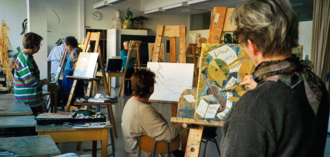 Henkilöitä maalaustelineiden edessä luokkahuoneessa. Pöydällä maalauksia.