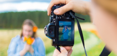 Two women are standing in a field. One takes a picture of the other with a camera.