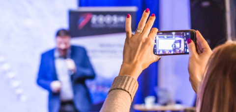 Taka-alalla näkyy lava, jolla on esiintyjä. Etualalla on henkilö kuvaamassa kännykällä lavaa.