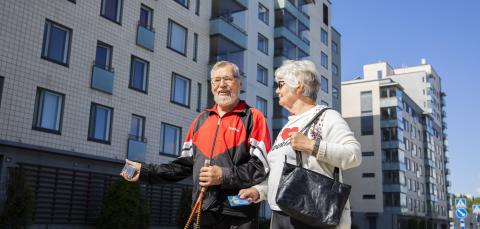 Ikääntynyt pariskunta viittaa bussia pysähtymään kerrostalon edessä. seisten.