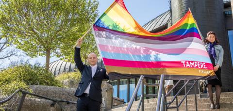 Kaksi henkilöä heiluttaa suurikokoista Tampereen omaa 13-raitaista pride-lippua pääkirjasto Metson rappusilla. 