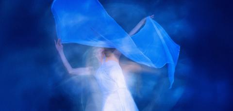 Valkoasuinen epätarkka hahmo tanssii vaalean kankaan kanssa sinistä taustaa vasten.