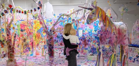 Nainen seisoo lapsi sylissään värikkäässä huoneessa, jossa valkoiselle taustalle on maalattu eri värein. Nainen ja lapsi katsovat suurta paperista tehtyä yksisarvista.