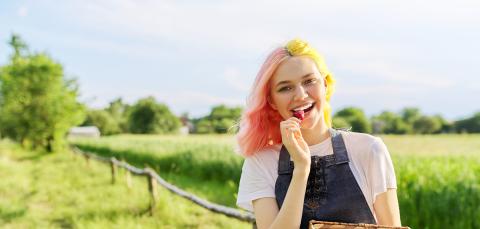 Henkilö kävelee pellon laidalla mansikkakori kädessään. Hän haukkaa yhtä mansikkaa.