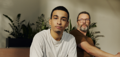 In the foreground sits a dark-haired man looking toward the viewer. Behind him sits an older bearded man.