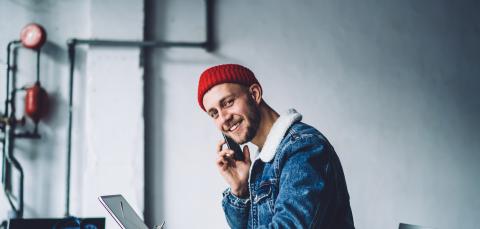 Nuori istuu kannettavan tietokoneen edessä ja soittaa puhelua. Hän hymyilee.