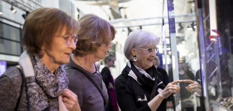 Three older women are viewing a museum exhibition together, the interior of the Vapriikki Museum Center is visible in the background.