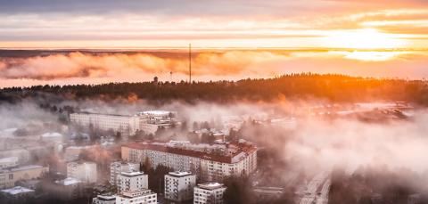 An aerial photograph taken from Näsinneula in Tampere, showing apartment buildings emerging through drifting mist. The sunset is a golden red.