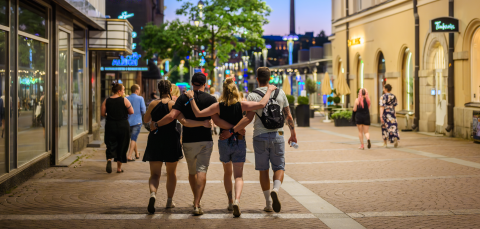 A group of four people walk arm in arm down a street illuminated by decorative lights, with shops and trees lining the pathway.