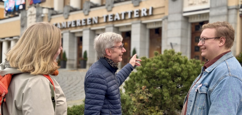 Nainen ja kaksi miestä seisoo Keskustorilla Tampereen teatterin edessä. Keskellä oleva harmaahiuksinen mies osoittaa kädellään teatteria.