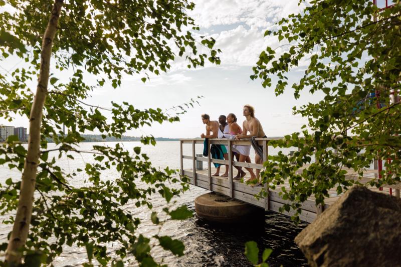 Four people are standing on a dock by the lake in the summer, with towels draped around them, looking out at the lake. In the foreground are trees with green leaves.