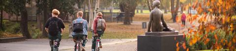Yksi henkilö kävelee ja kaksi pyöräilee syksyisessä kaupunkipuistossa.