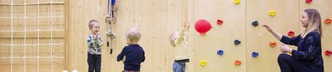 Liikuntasalissa yksi aikuinen ja kolme lasta, joista yksi heittää ilmapalloa. Taustalla kiipeilyseinä.