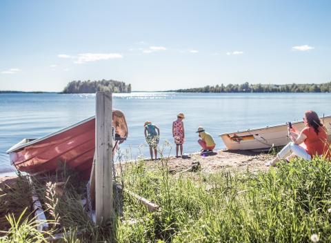On the shore of a lake, three children explore the sand, a person sits on the edge of the grass with a phone in hand, two boats on docks.