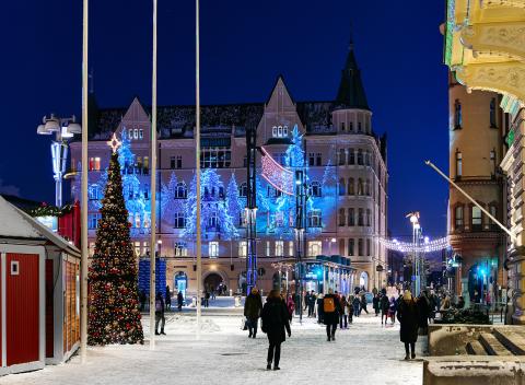 Ihmisiä kävelee lumisella Keskustorilla, taustalla valotaiteella valaistu jugend-rakennus ja etualalla osa Joulutorista sekä Raatihuoneen kulmaa.