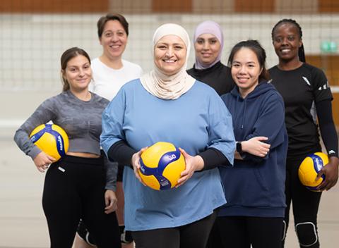 Six women on a volleyball field. All of them smiling to a camera. 