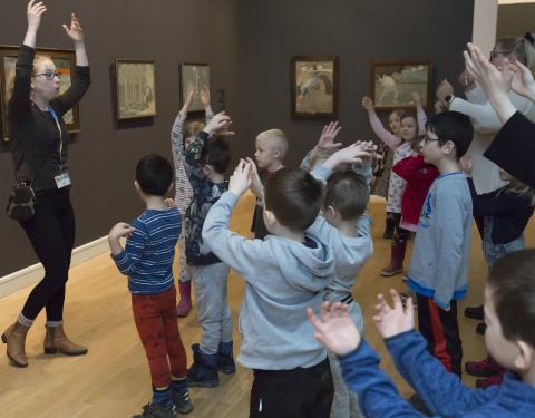 Lapsiryhmä ja opas loruleikeissä kädet ylhäällä taidenäyttelyssä.