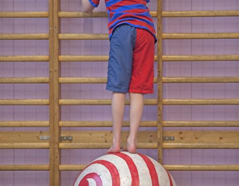 Lapsi tasapainoilee suuren pallon päällä värikkäässä asussa.