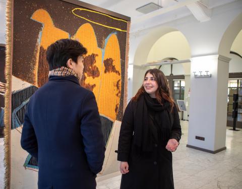 Suuressa galleriatilassa roikkuu lattiasta kattoon ulottovia ryijyä. Niiden edessä kaksi henkilöä juttelevat keskenään, toinen on katsojaan selin, toinen hymyilee ystävälleen.