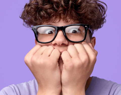 Nuori kiharatukkainen poika on innostuneen näköinen. Hän pitää käsiään kasvojen edessä ja katsoo kameraan silmälasiensa takaa.