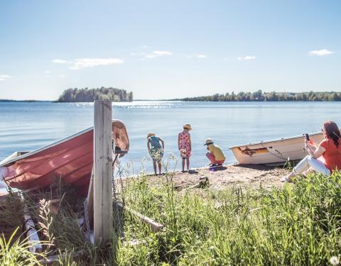 On the shore of a lake, three children explore the sand, a person sits on the edge of the grass with a phone in hand, two boats on docks.