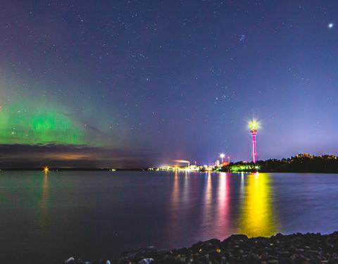 A night view from Lake Näsijärvi, green northern lights in the sky, the Näsinneula and the city lights shining in the distance.