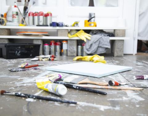 Pensseleitä, maalauspohja, maalituubeja sekä muita taiteilijan tarvikkeita lattialla työhuoneesssa.