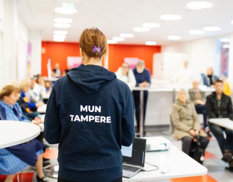 A city employee wearing a Mun Tampere hoodie is in front of the audience talking to them.
