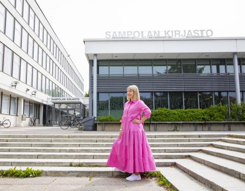 Henkilö seisoo vaaleanpunaisessa pitkässä mekossa rakennuksen edessä, jossa lukee Sampolan kirjasto.