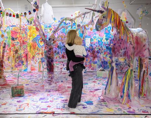 Nainen seisoo lapsi sylissään värikkäässä huoneessa, jossa valkoiselle taustalle on maalattu eri värein. Nainen ja lapsi katsovat suurta paperista tehtyä yksisarvista.