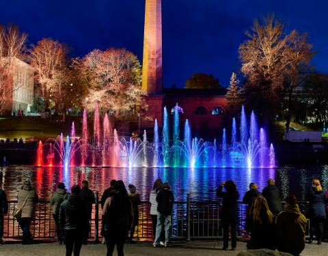 Colourful fountains at the Dancing Fountains show in Tammerkoski rapids at dusk. People watch the show from the railing.