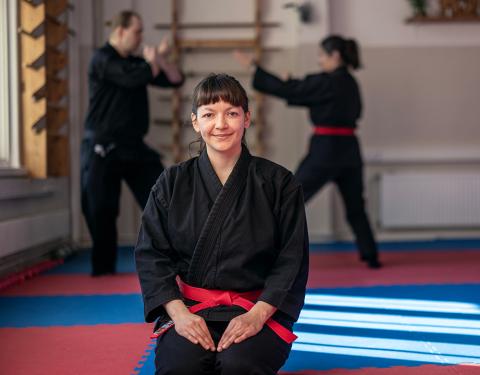 A woman is looking at the camera in a martial arts suit with two people practising martial arts at the backgorund