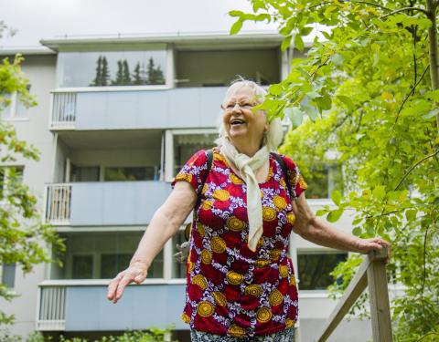 Ikääntynyt nainen hymyilee vehreässä kesämaisemassa kerrostalo taustalla