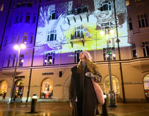 A person stands on the Jugend Square looking up to the left, with a Moomin-themed lightwork reflected on the building's façade in the background.