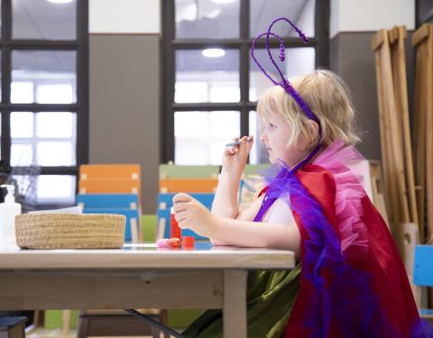 Lapsi istuu pöydän ääressä värikkäissä vaatteissa askartelemassa. Päässä on hiuspanta, jossa on tuntosarvet.