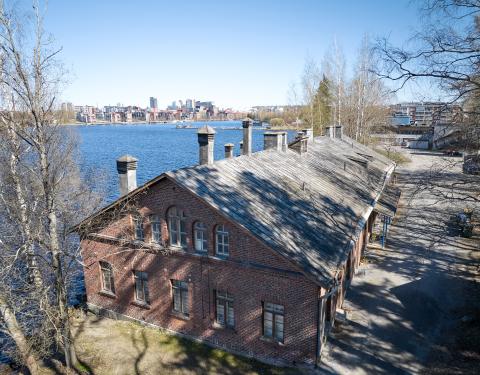 Ilmakuvan etualalla kuva punatiilisestä pesularakennuksesta. Taustalla näkyy Pyhäjärveä ja keskustan siluetti.