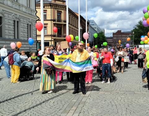 Vammaisten kulkue kävelee Keskustorilla. Etummaiset pitelevät käsissään pride-lippua. Monilla on värikkäitä ilmapalloja. Osallistujissa näkyy muun muassa pyörätuolilla kulkevia.