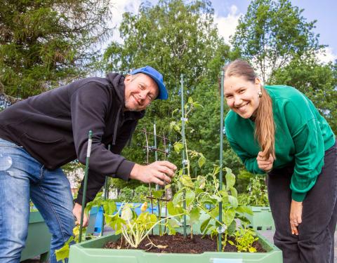 Two people, with a planter box between them, are looking at the camera. In the background are leafy green trees and planter boxes.