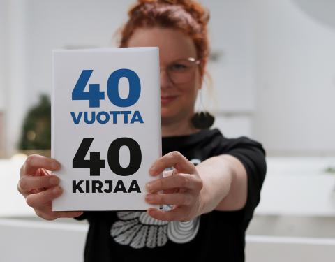 Tuija seisoo Metson parvikerroksessa ja pitää osittain kasvojensa edessä kirjaa, jonka kannessa on teksti 40 vuotta, 40 kirjaa. 