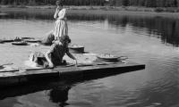 Yksi nainen pesee mattoa järvellä. Toinen katselee taustalla.
